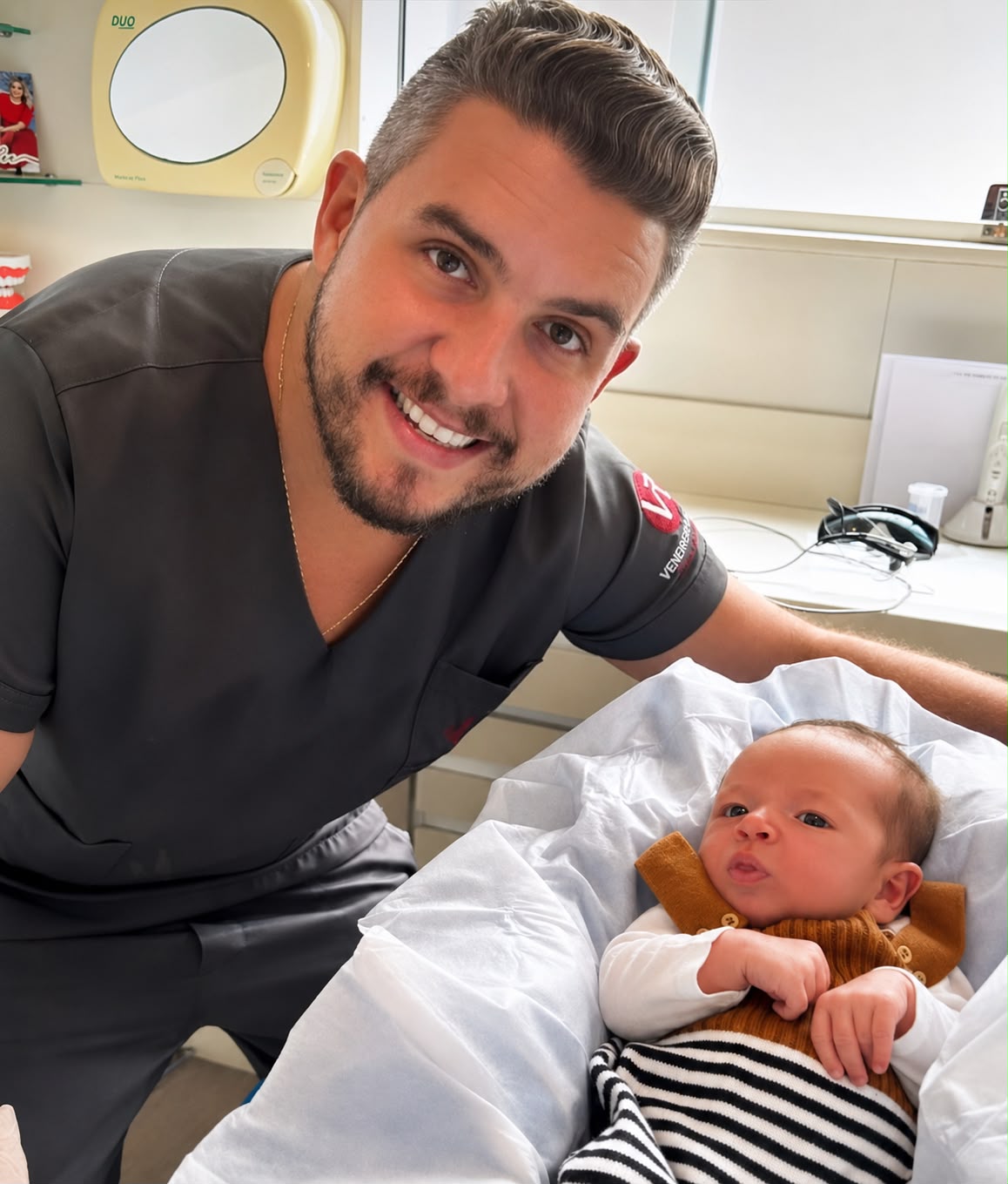 Dr. Henrique Veneroso sorrindo ao lado de um recém-nascido deitado na maca do consultório